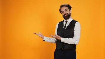 Skilled classy butler pointing aside and working at five star restaurant. Young professional valet dressed in suit and tie showing direction on left or right sides of yellow studio. photo