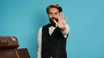 Doorman raises hand to show stop sign against blue background, portraying advertisement and expressing refusal symbol in studio. Bellboy hotel porter presenting his rejection, luggage bags. photo