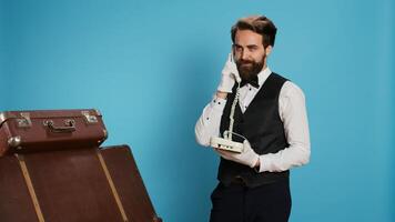 Classy bellhop using landline phone on camera, talking to guests about booking accommodation. Skilled professional porter making reservations remotely with office cord telephone. photo