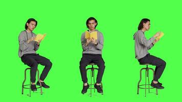 Trendy person reading novel book on chair against full body greenscreen template, enjoying old school lecture hobby. Young asian adult holding literature or fiction book to read in studio. video