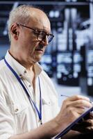 Aged computer scientist in crypto mining farm with specialized infrastructure using Blockchain technology. Fintech employee holding clipboard, inspecting asic racks solving complex hashing operations photo