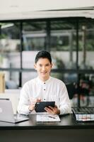 Young handsome man typing on tablet and laptop while sitting at the working tabl in office photo