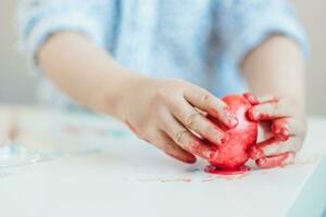 un niño pone un rojo Pascua de Resurrección huevo en un estar con su manos manchado con pintar en un blanco mesa. foto