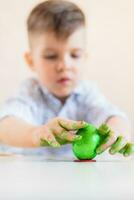 A boy puts a green Easter egg on a stand with his hands stained with paint on a white table. photo