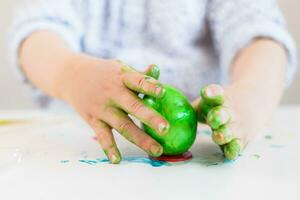 A child puts a green Easter egg on a stand with his hands stained with paint on a white table. photo