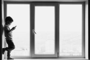 A little boy in a medical mask is sitting on the window of the house in quarantine with a phone in his hands.Prevention of coronavirus and Covid - 19 photo