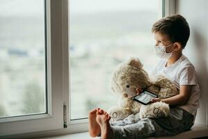 A little boy sits on a window with a teddy bear in quarantine and plays in a mobile phone. Prevention of coronavirus and Covid - 19 photo