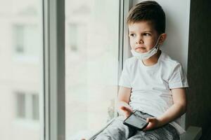 un pequeño niño en un médico máscara se sienta en cuarentena a hogar en un ventana con un teléfono en su manos.prevención de coronavirus y codicioso - 19 foto