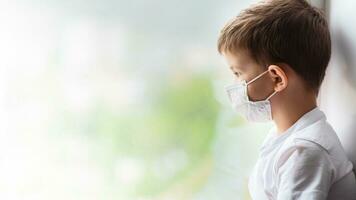 a child in a medical mask is sitting at home in quarantine because of coronavirus and covid -19 and looks out the window. photo