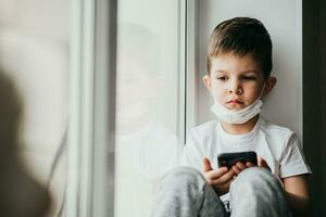 A small child in a medical mask sits quarantined at home on a window with a phone in his hands.Prevention of coronavirus and Covid - 19 photo