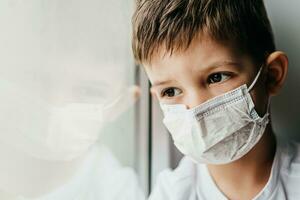 un niño en un médico máscara es sentado a hogar en cuarentena porque de coronavirus y codicioso -19 y mira fuera el ventana. foto
