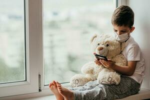 A little boy sits on a window with a teddy bear in quarantine and plays in a mobile phone. Prevention of coronavirus and Covid - 19 photo