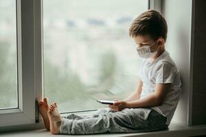 A small child in a medical mask sits quarantined at home on a window with a phone in his hands.Prevention of coronavirus and Covid - 19 photo