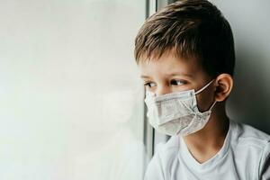 un pequeño chico en un médico máscara es sentado a hogar en cuarentena porque de coronavirus y codicioso -19 y mira fuera el ventana. foto