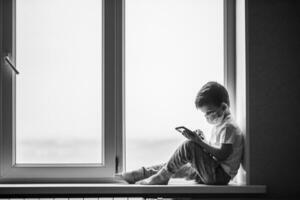 un pequeño chico en un médico máscara es sentado en el ventana de el casa en cuarentena con un teléfono en su manos.prevención de coronavirus y codicioso - 19 foto