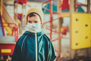 un niño en un médico máscara soportes cerca el patio de recreo durante el pandemia de coronavirus y codicioso - 19 foto