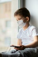 A small child in a medical mask sits quarantined at home on a window with a phone in his hands.Prevention of coronavirus and Covid - 19 photo