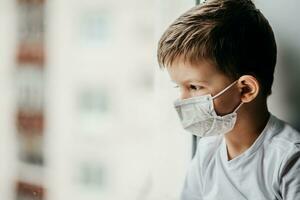 a child in a medical mask is sitting at home in quarantine because of coronavirus and covid -19 and looks out the window. photo