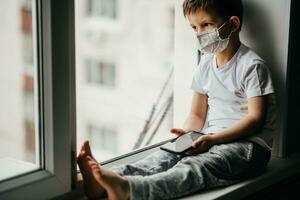 A little boy in a medical mask is sitting on the window of the house in quarantine with a phone in his hands.Prevention of coronavirus and Covid - 19 photo