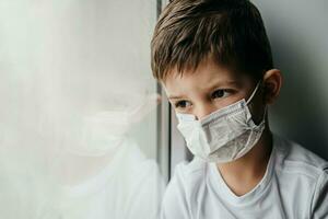 un pequeño chico en un médico máscara es sentado a hogar en cuarentena porque de coronavirus y codicioso -19 y mira fuera el ventana. foto