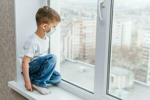 a child in a medical mask is sitting at home in quarantine because of coronavirus and covid -19 and looks out the window. photo