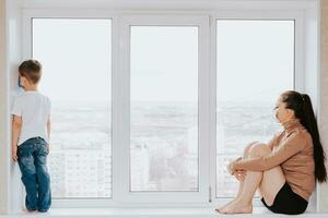 A woman with a child in medical masks is sitting on the window at home in quarantine and looking out the window. Prevention of Coronovirus and Covid -19 photo