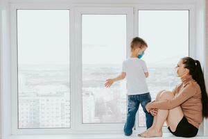 A woman with a child in medical masks is sitting on the window at home in quarantine and looking out the window. Prevention of Coronovirus and Covid -19 photo