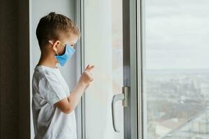 a child in a medical mask is sitting at home in quarantine because of coronavirus and covid -19 and looks out the window. photo