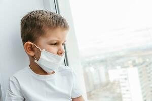 un niño en un médico máscara es sentado a hogar en cuarentena porque de coronavirus y codicioso -19 y mira fuera el ventana. foto