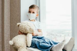a small child in a mask sits at home in quarantine and looks out the window in a place with a teddy bear. Prevention of Coronavirus and Covid - 19. Concept photo