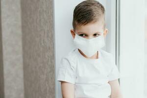 a child in a medical mask is sitting at home in quarantine because of coronavirus and covid -19 and looks out the window. photo