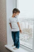 a child in a medical mask is sitting at home in quarantine because of coronavirus and covid -19 and looks out the window. photo