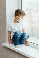 a child in a medical mask is sitting at home in quarantine because of coronavirus and covid -19 and looks out the window. photo