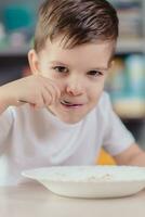 Beautiful boy eats milk porridge. Cute baby having breakfast sitting at a table in the kitchen at home. photo