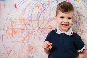 A cheerful boy with a pencil stands near the wall that he painted. A child is engaged in creativity at home. photo