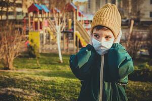 un niño en un médico máscara soportes cerca el patio de recreo durante el pandemia de coronavirus y codicioso - 19 foto