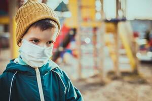 un niño en un médico máscara soportes cerca el patio de recreo durante el pandemia de coronavirus y codicioso - 19 foto