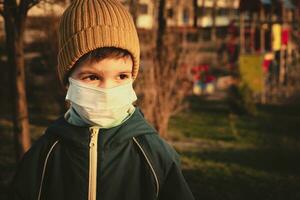 un niño en un médico máscara soportes cerca el patio de recreo durante el pandemia de coronavirus y codicioso - 19 foto