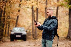 el hombre viajes por coche. el chico en el teléfono mira a dónde él es en el navegador. caminante perdido en el otoño bosque concepto. foto