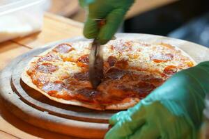 A person wearing green gloves is cutting thin-crust, small-size pizzas on a wooden tray with a pizza cutter. photo