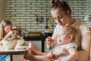 Two young mothers together feed their happy babies milk porridge in the kitchen photo