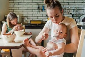 Two young mothers together feed their happy babies milk porridge in the kitchen photo