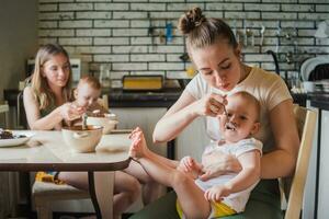 Two young mothers together feed their happy babies milk porridge in the kitchen photo