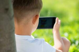 A beautiful child sits on the grass and looks at the phone in the summer at sunset. Cute boy with mobile in hand photo