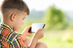 el niño mira dentro el móvil mientras sentado en el verano en el calle. pequeño niño con un teléfono en su manos. foto