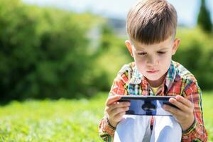 Cheerful child sitting on the grass looks cartoons in the phone in the summer at sunset. Cute boy having fun in nature photo