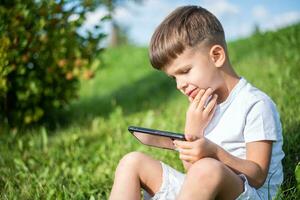 alegre niño sentado en el césped mira dibujos animados en el teléfono en el verano a puesta de sol. linda chico teniendo divertido en naturaleza foto