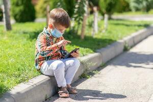 A child in a medical mask sits on the grass and looks at the phone in the summer at sunset. Boy with a mobile phone in his hand. Prevention against coronavirus Covid-19 during a pandemic photo