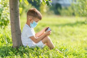 A kid in a medical mask sits on the grass and looks in the phone cartoons in the summer at sunset. Child with a mobile phone in his hands. Prevention against coronavirus Covid-19 during a pandemic photo