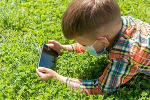 A kid in a medical mask lies on the grass and looks in the phone cartoons in the summer at sunset. Child with a mobile phone in his hands. Prevention against coronavirus Covid-19 during a pandemic photo
